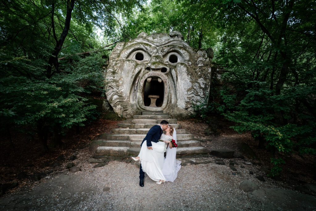 Scatto di un reportage matrimoniale nel Bosco Sacro di Bomarzo, conosciuto meglio come Parco dei Mostri. Lo sposo prende la sposa, la cinge a sè e durante un casquet, e insieme entrano nel campo visibo della bocca dell'inferno